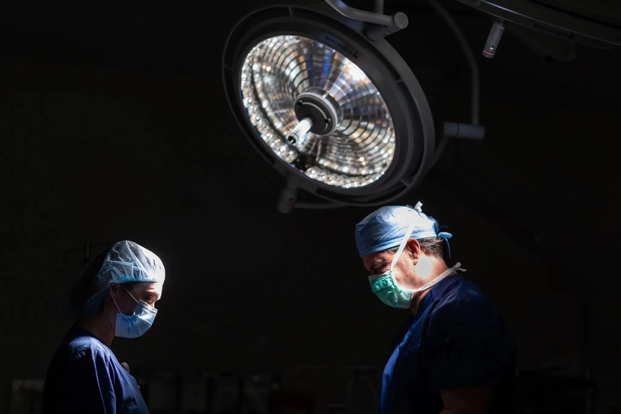 Two surgeons wearing masks and surgical caps working under a bright operating room light.