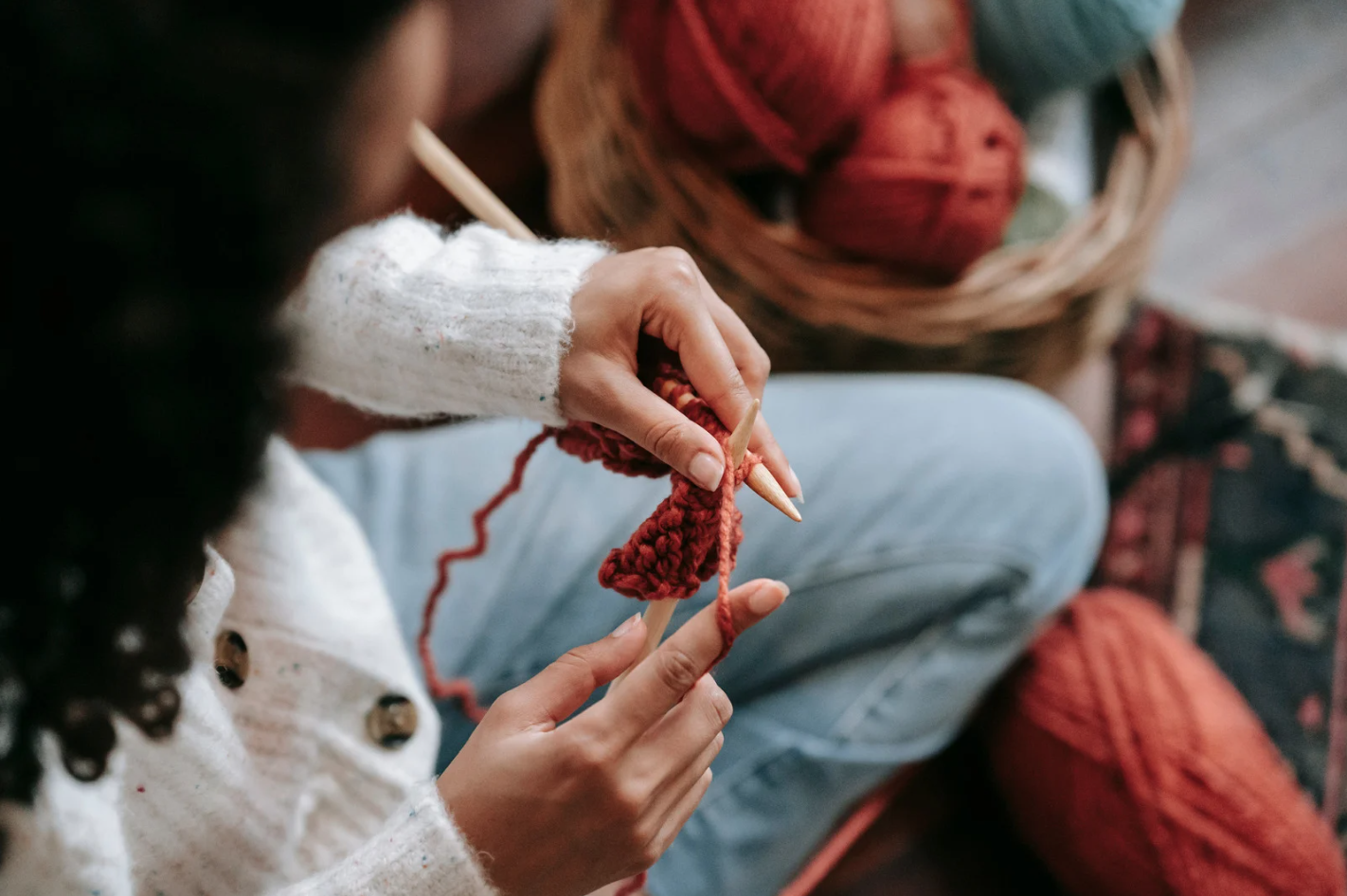 Close-up of a person knitting with red yarn, holding wooden knitting needles beside a basket of yarn balls.