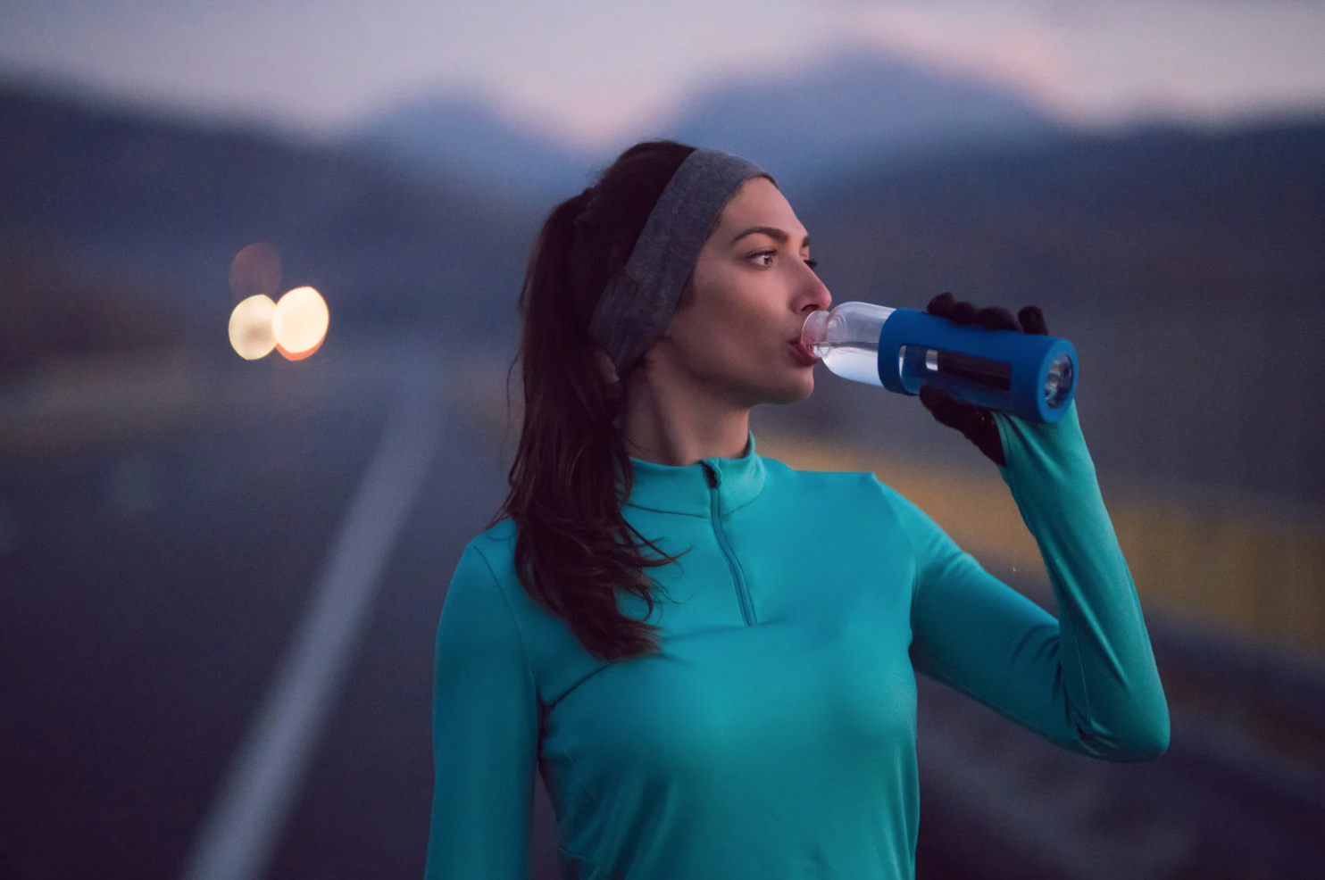 Woman in athletic wear drinking water from a bottle while standing on a road at dusk.