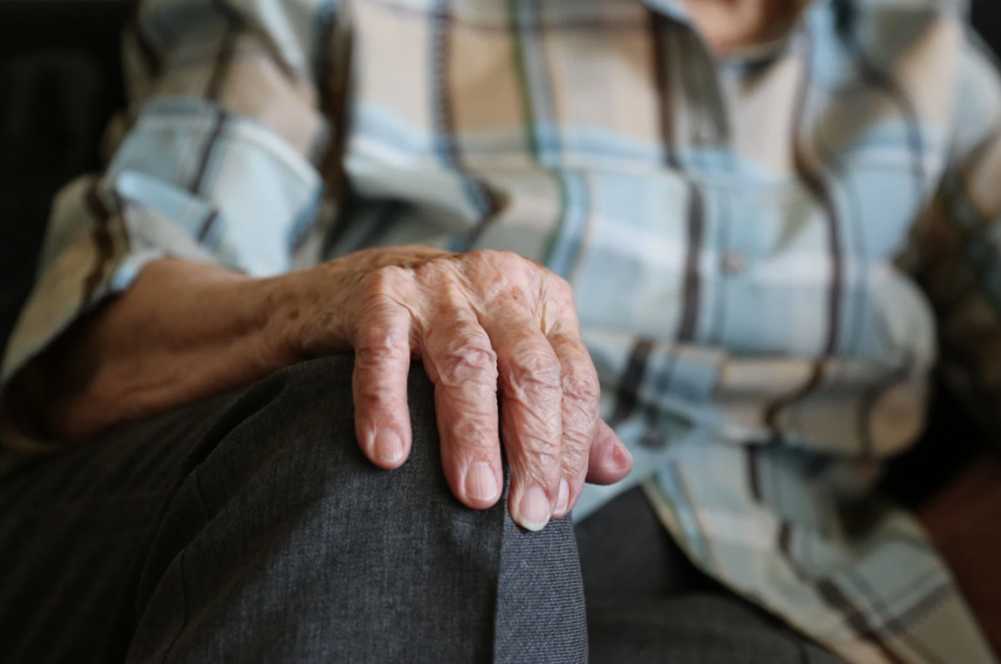 Close-up of an elderly person’s hand resting on their knee, wearing a plaid shirt and gray pants.