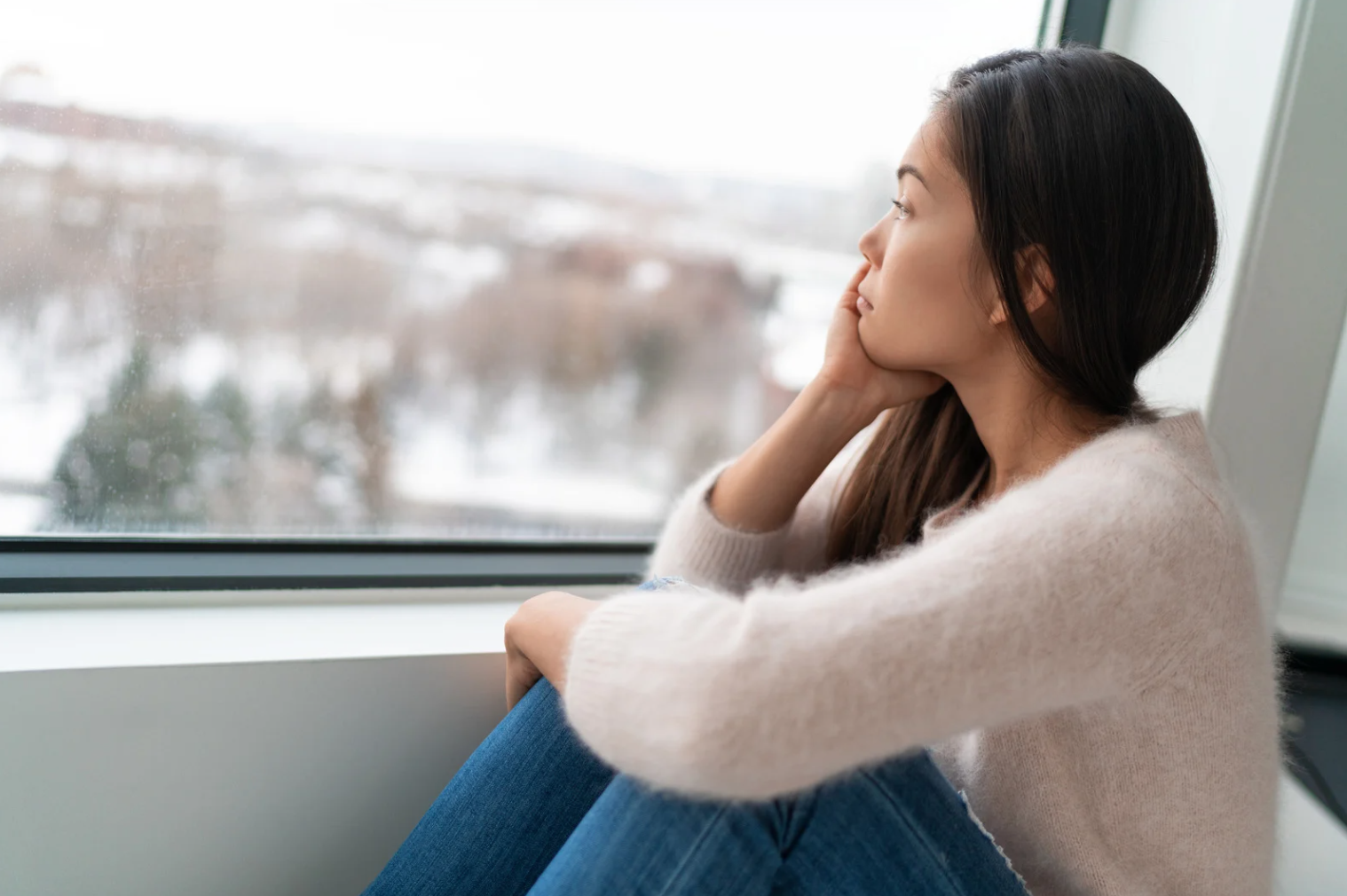Young woman sitting by a window on a winter day, resting her chin on her hand while looking outside thoughtfully.