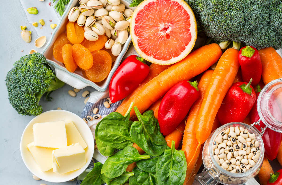 Assortment of healthy foods including broccoli, carrots, spinach, red peppers, grapefruit, pistachios, dried apricots, beans, and butter on a table.