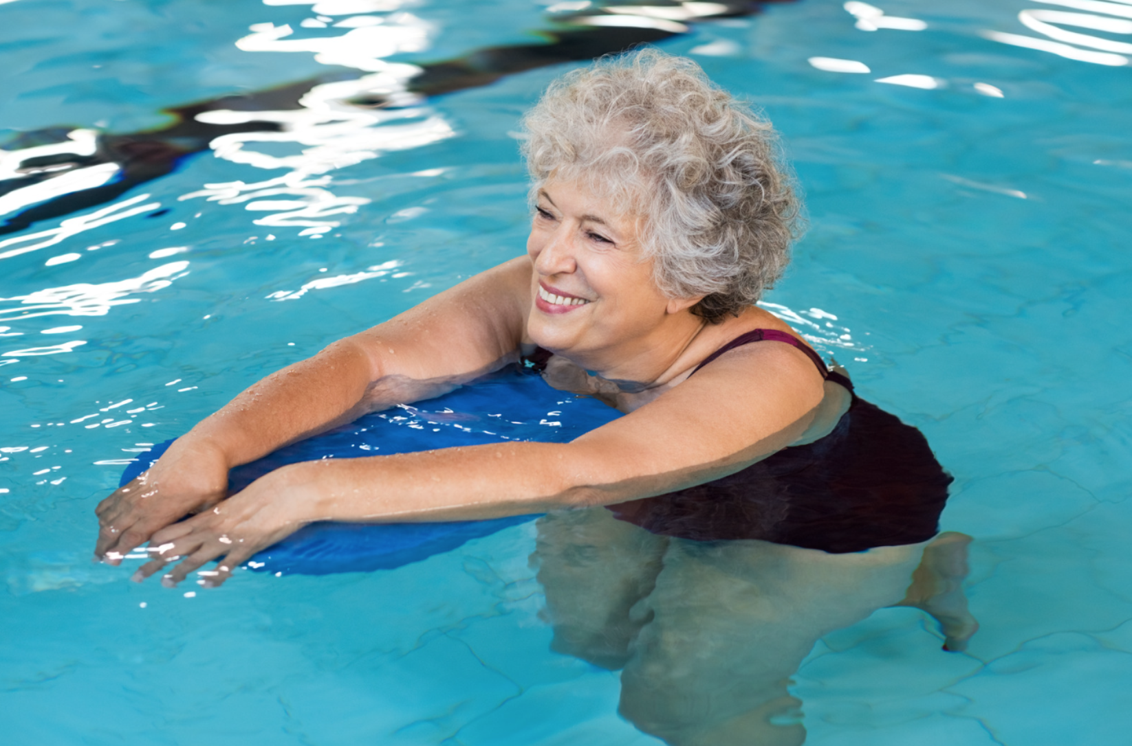 Smiling older woman in a swimsuit using a kickboard while exercising in a swimming pool.