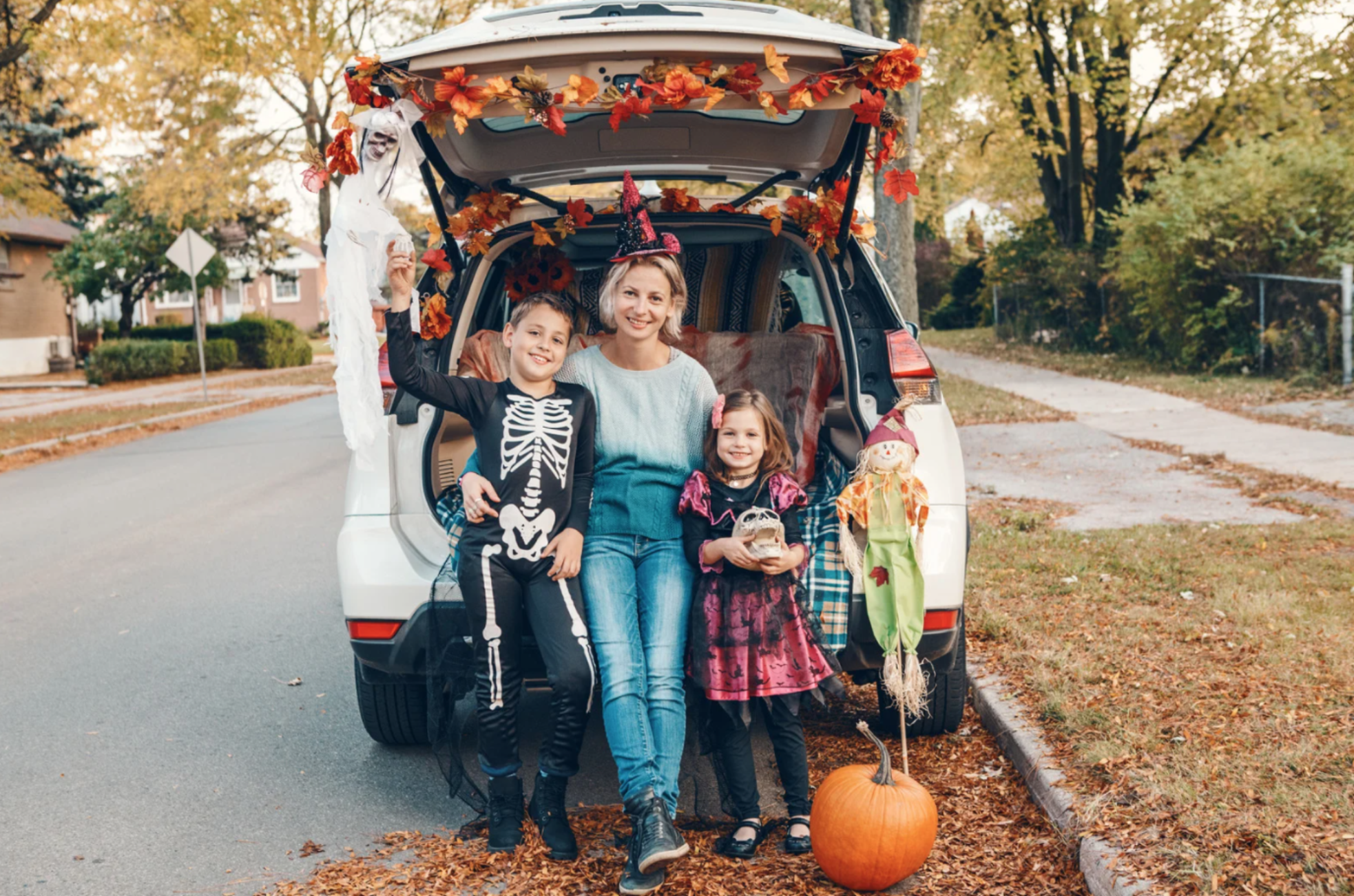 Mother with two children in Halloween costumes posing by a decorated car trunk with autumn leaves, a pumpkin, and festive decorations during a trunk-or-treat event.